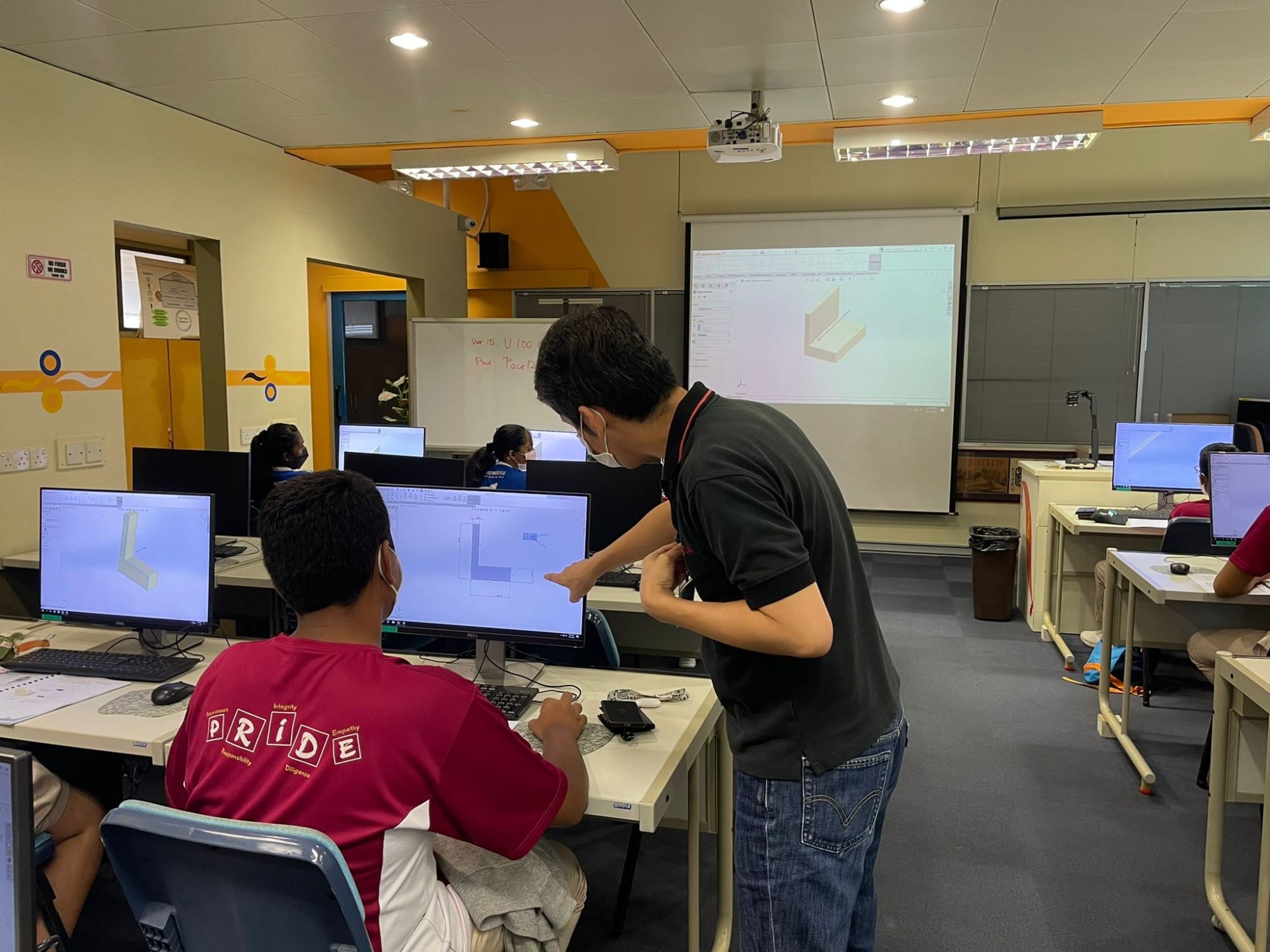 A teacher points at a student's monitor showing CAD software in a computer lab with a projected 3D model on screen.